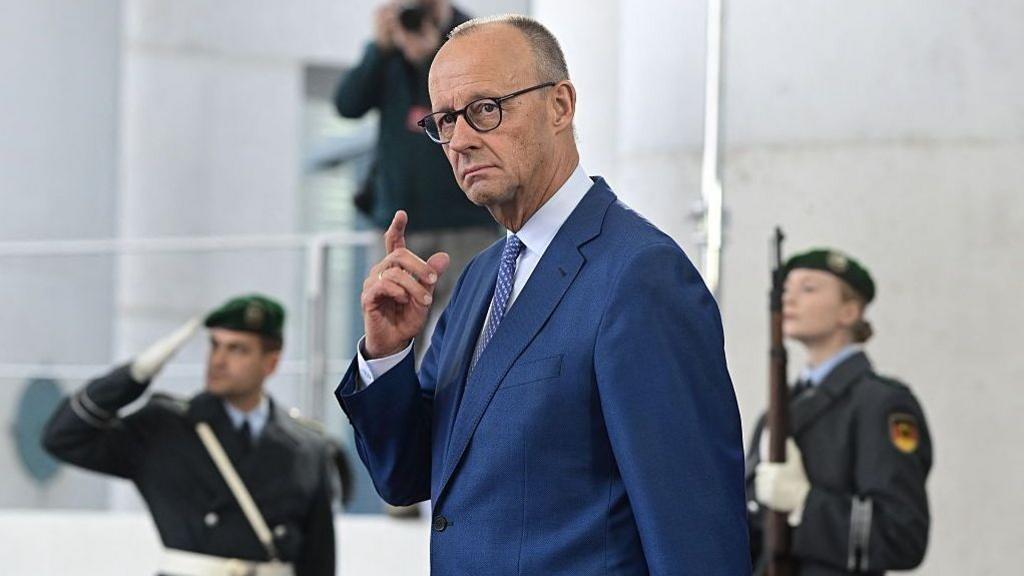 German Chancellor Friedrich Merz in a blue suit with hand raised, two military officers stand behind him